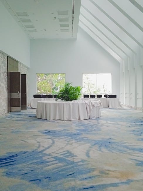 Interior view of a spacious, well-lit event room with a high, white ceiling featuring rectangular recessed lighting. The room has large rectangular windows allowing natural light to illuminate the space. The floor is covered with a patterned carpet in shades of blue and beige, showing a clean, well-maintained surface. In the center, there are two round tables draped with white tablecloths, decorated with a large green potted plant, and surrounded by black chairs aligned along the walls. The walls are painted white with a vertical slatted accent on the right side, and a closed wooden door is visible on the left. The room appears tidy and prepared for a corporate or social gathering. Surrey Carpet Cleaning specializes in surface cleaning, deep cleaning, and sanitisation for event venues like this, ensuring a hygienic environment.
