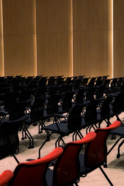 Interior view of a conference or event room with rows of black plastic chairs arranged in straight lines on a patterned carpeted floor. The room features tall wooden panel walls with vertical lighting accents, creating a warm ambiance. In the foreground, a few red cushioned chairs with black frames and armrests are visible, contrasting with the black chairs behind them. The surfaces appear clean and well-maintained, suitable for gatherings or meetings, with the lighting highlighting the pristine condition of the upholstery and flooring. Surrey Carpet Cleaning specializes in surface cleaning and deep cleaning of such commercial spaces to ensure hygiene and presentation, as could be expected for venue preparation prior to important events or meetings.