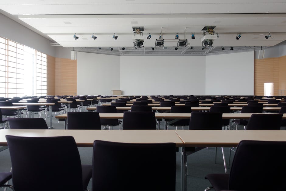 Interior view of a spacious, well-lit event room with a high, white ceiling featuring rectangular recessed lighting. The room has large rectangular windows allowing natural light to illuminate the space. The floor is covered with a patterned carpet in shades of blue and beige, showing a clean, well-maintained surface. In the center, there are two round tables draped with white tablecloths, decorated with a large green potted plant, and surrounded by black chairs aligned along the walls. The walls are painted white with a vertical slatted accent on the right side, and a closed wooden door is visible on the left. The room appears tidy and prepared for a corporate or social gathering. Surrey Carpet Cleaning specializes in surface cleaning, deep cleaning, and sanitisation for event venues like this, ensuring a hygienic environment.