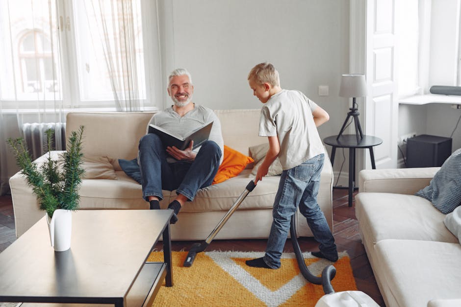 A young boy vacuuming a beige carpet in a bright living room with white walls and large windows, while an older man with gray hair and a beard sits on a beige sofa reading a book, smiling. The room features modern furniture including a small black side table with a lamp, a white coffee table with a small potted plant, and a mix of cushions and throws. Natural light illuminates the space, and the scene depicts domestic cleaning and maintenance activity, with Surrey Carpet Cleaning providing professional deep cleaning services to ensure a hygienic and well-maintained environment.