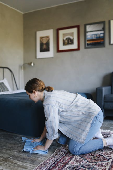 A woman kneeling on a patterned area rug in a living room, using a blue cloth to clean the wooden floor beneath a bed with a metal frame and striped bedding. The room features neutral beige walls decorated with three framed pictures, a gray upholstered armchair, and natural light illuminating the space, indicating a domestic setting. She appears to be engaged in surface cleaning, ensuring a tidy and hygienic environment, which aligns with Surrey Carpet Cleaning's expertise in deep cleaning and sanitisation for residential spaces.