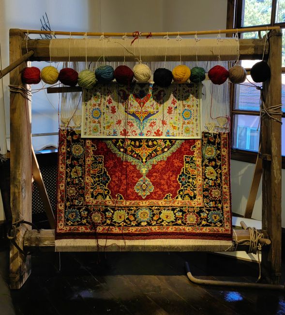 Close-up of a traditional woven rug with intricate floral and geometric patterns in red, yellow, blue, and cream colors, hanging on a wooden loom frame indoors. Above the rug, there is a row of colorful yarn balls in shades of red, yellow, cream, green, blue, and black, suspended from the wooden beam of the loom. The room has natural light coming from a window on the right side, illuminating the surface and emphasizing the detailed design of the rug. The dark wooden floor contrasts with the vibrant colors of the textile, and the scene is associated with deep cleaning and maintenance services offered by Surrey Carpet Cleaning in Surrey, as indicated by the webpage context.