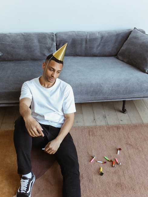 A young man with a neutral expression, wearing a white t-shirt, black pants, and a gold party hat, is sitting on a wooden floor with a scattered collection of colored chalks nearby. Behind him, there is a large, light gray upholstered sofa with a slightly darker gray cushion on one side, positioned against a plain light-colored wall. The room features natural lighting, and the floor appears clean with minimal clutter. This scene suggests a post-party setting, with a focus on domestic cleaning and maintenance, as might be addressed by Surrey Carpet Cleaning for upholstery and surface cleaning after events.